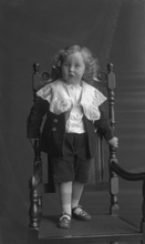 Male child with long blonde ringlets standing on a chair wearing a Little Lord Fauntleroy suit.#