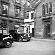Macrae & Dick Taxi Booking Office in Station Square, the site now occupied by Mail Boxes Etc., with Ben Wyvis Kilts upstairs. The train station building is on the right. On the left are the offices of McGruther & Marshall Ltd, coal and cement merchants and shipping agents. *