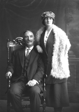 Couple, he seated wearing suit, she standing wearing hat and fur shawl. #