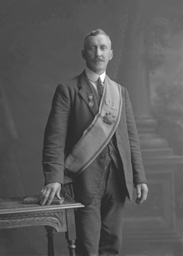 Suited man wearing over shoulder sash featuring the letters 'C' and 'L', alongside two different large medals.#