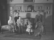 Family with three children and labrador dog seated in lounge room in front of fireplace. Man wearing kilt.#