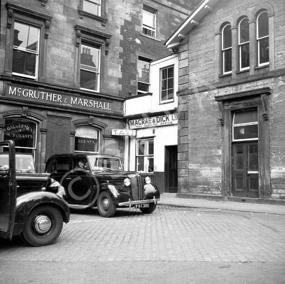 Macrae & Dick Taxi Booking Office in Station Square, the site now occupied by Mail Boxes Etc., with Ben Wyvis Kilts upstairs. The train station building is on the right. On the left are the offices of McGruther & Marshall Ltd, coal and cement merchants and shipping agents. *