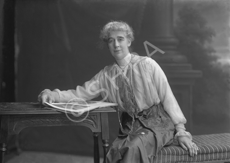 Elderly woman leaning on desk reading a book, wearing satin skirt with timepiece on a long cord.#