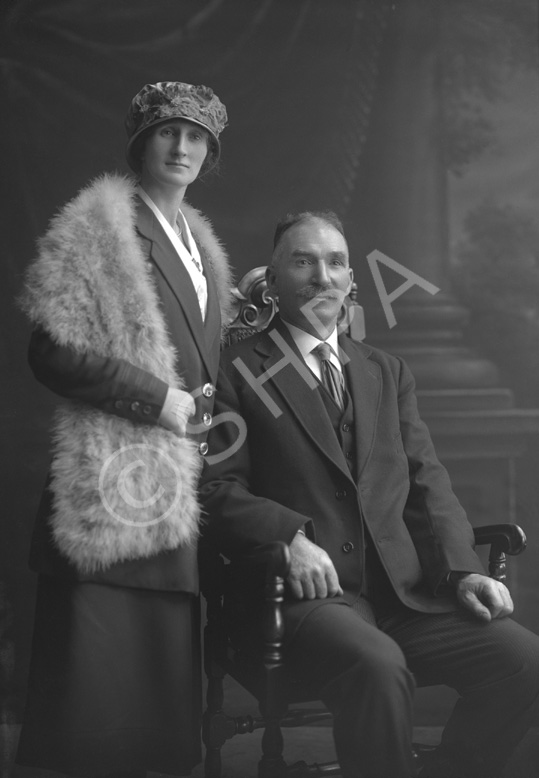Couple, he seated wearing suit, she standing wearing hat and fur shawl.#