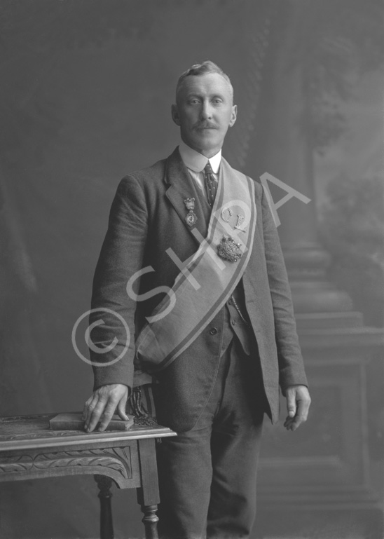 Suited man wearing over shoulder sash featuring the letters 'C' and 'L', alongside two different large medals.#